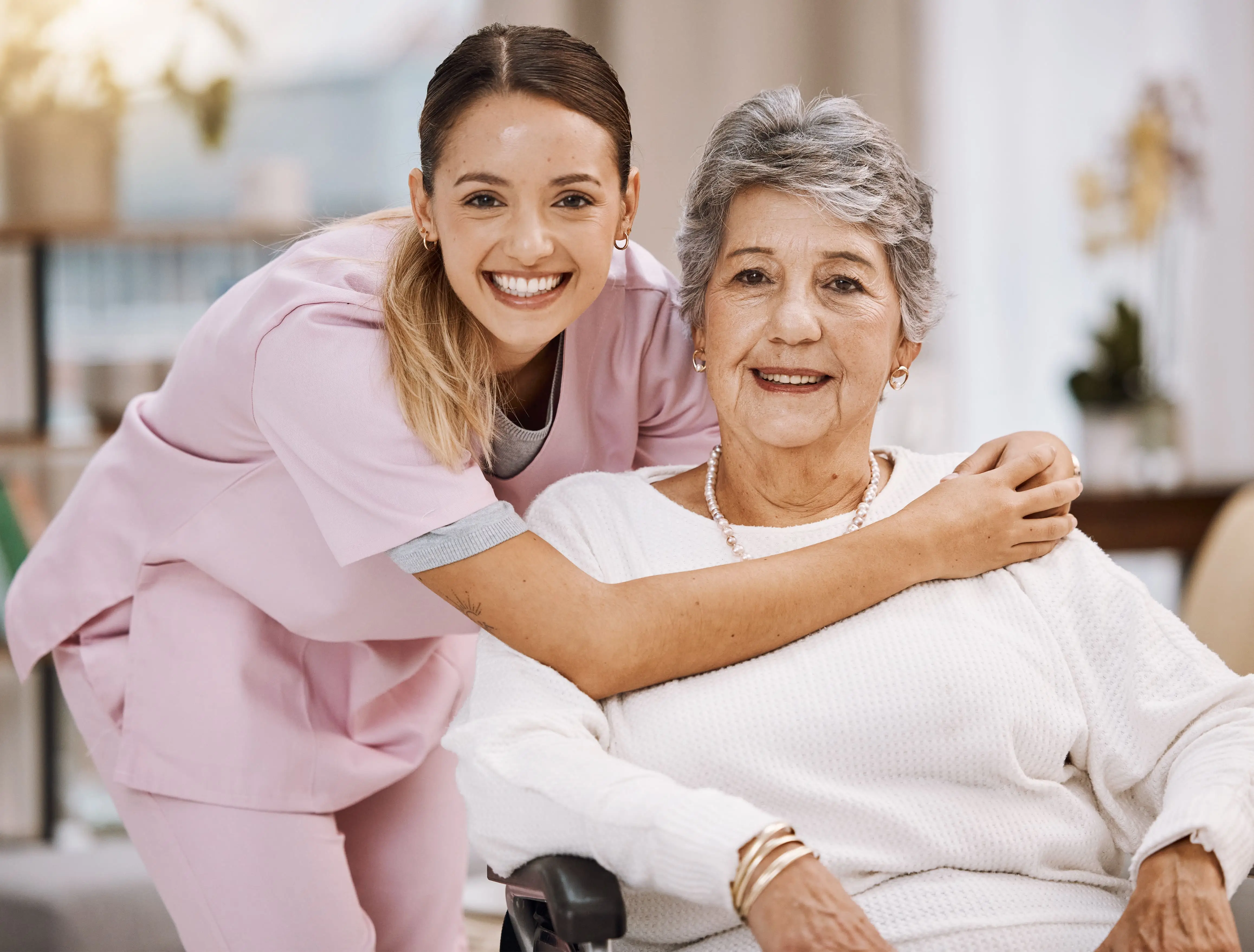 Smiling OC HomeCare caregiver in pink scrubs embracing a happy elderly woman in a wheelchair, representing compassionate in-home care in Orange County, CA